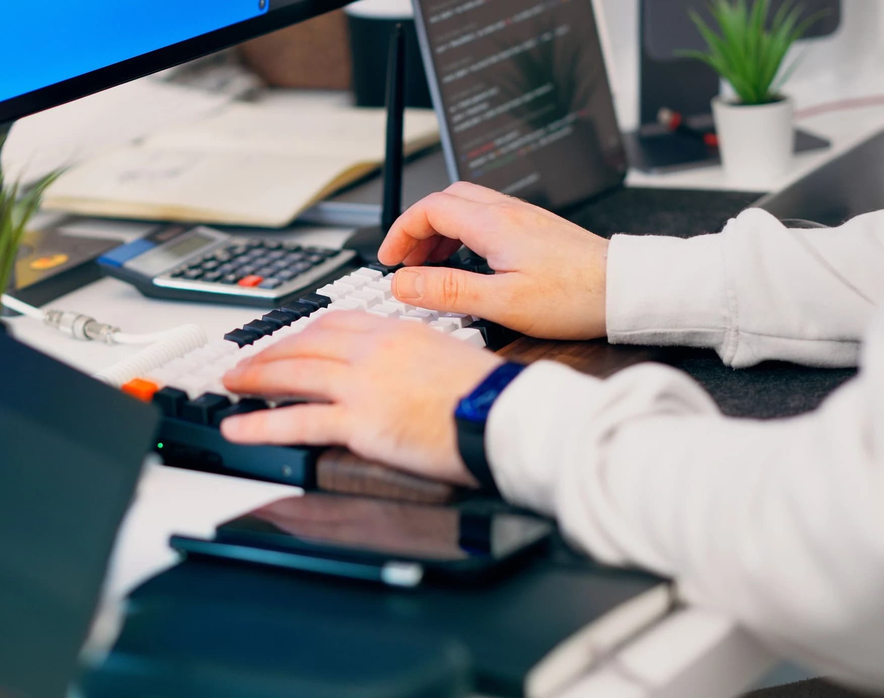 Handen typen op een toetsenbord aan een bureau met twee schermen.
